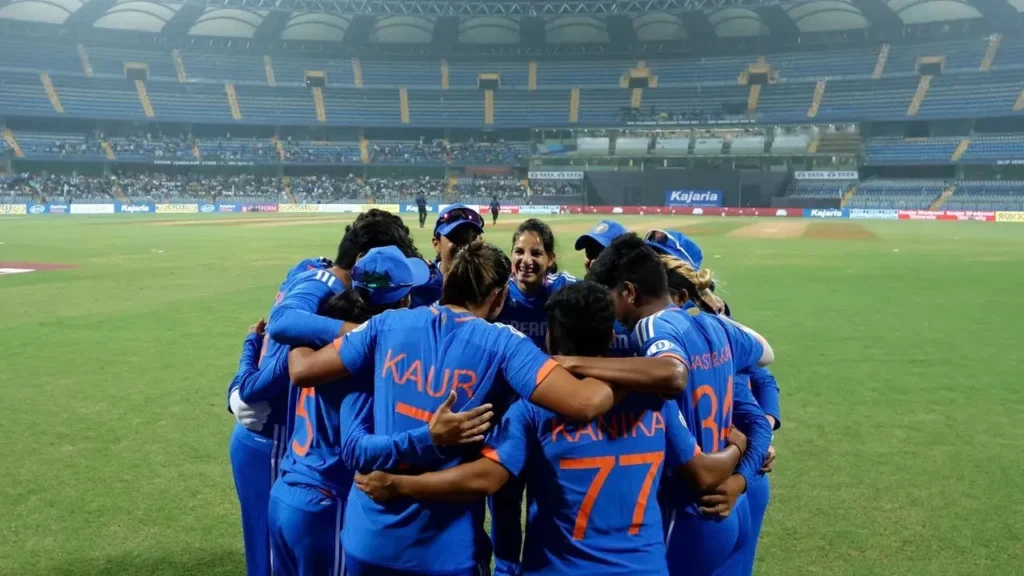 India vs Australia women's cricket match, with a player from the Indian team in a blue jersey batting against an Australian bowler in a yellow jersey, on a sunny day at a cricket stadium.