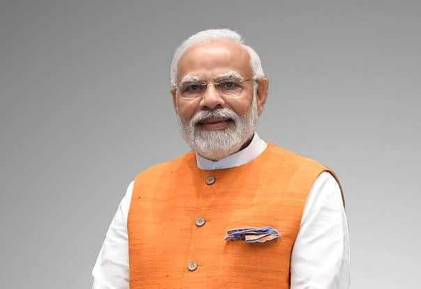 Prime Minister Narendra Modi standing on a stage, addressing a large, enthusiastic crowd with a backdrop of a development project under construction.