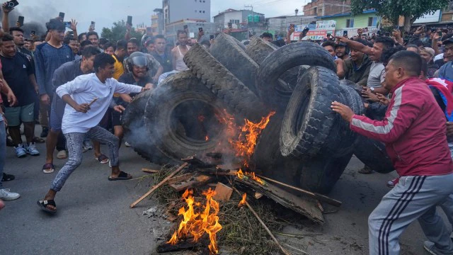 A nighttime scene of Nepalese Gen Z protesters holding "Stop Corruption" banners with a building on fire in the background, signifying the intensity of the unrest.

